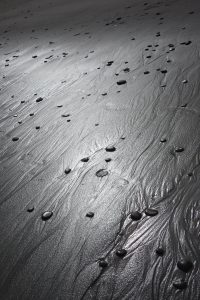 A photograph of a beach of black sand, dotted with smooth black pebbles.