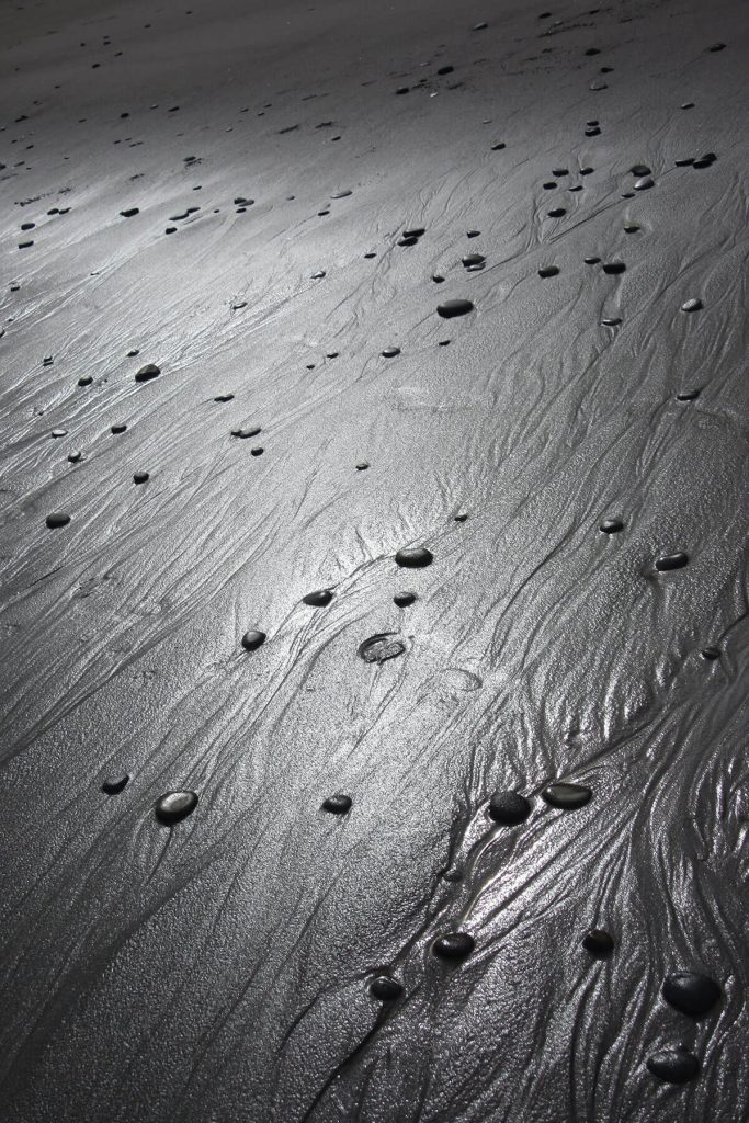A photograph of a beach of black sand, dotted with smooth black pebbles.