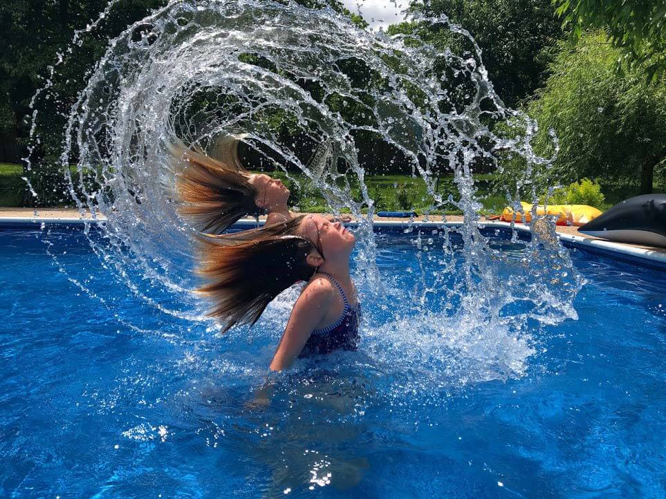 A photograph of twin girls in a pool flicking their water-soaked hair in an arc.