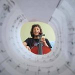 A photograph of a girl holding a violin, seen through a tube of music sheets.