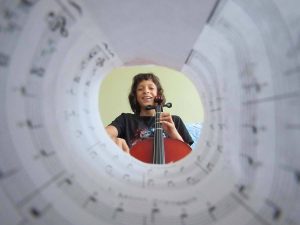A photograph of a girl holding a violin, seen through a tube of music sheets.